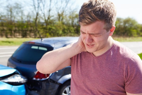 A man holds his neck in pain after a car accident, standing near two damaged vehicles. - Auto Accident Chiropractor