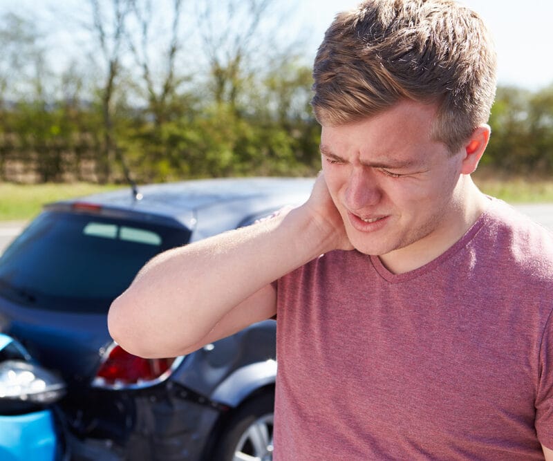 A man holds his neck in pain after a car accident, standing near two damaged vehicles. - Auto Accident Chiropractor