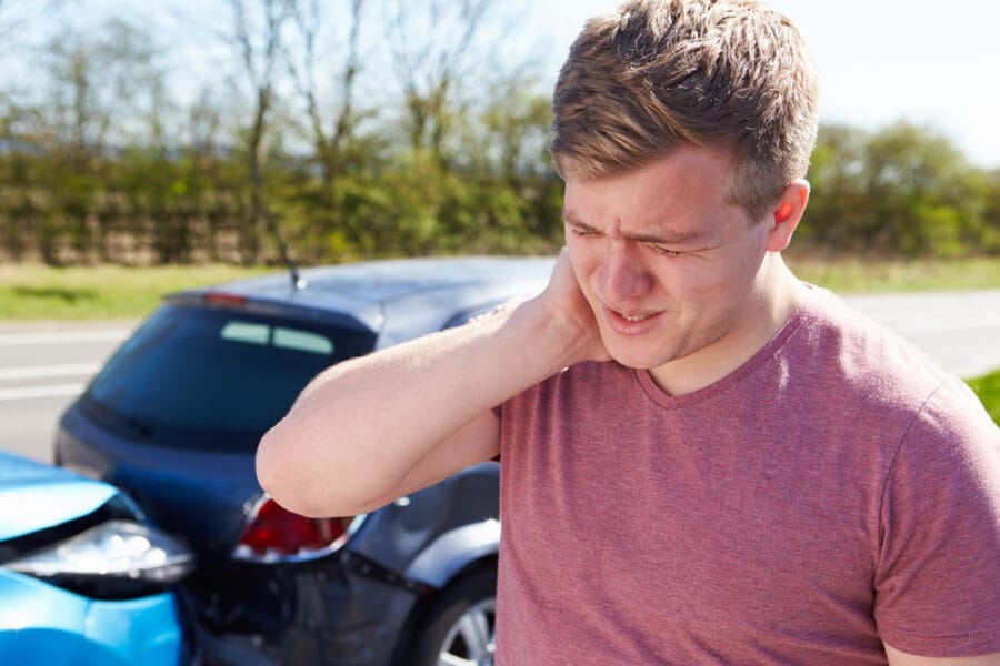 A man holds his neck in pain after a car accident, standing near two damaged vehicles. - Auto Accident Chiropractor